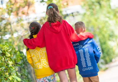 Three children in colorful hoodies walking along a tree-lined path, enjoying a sunny day outdoors.