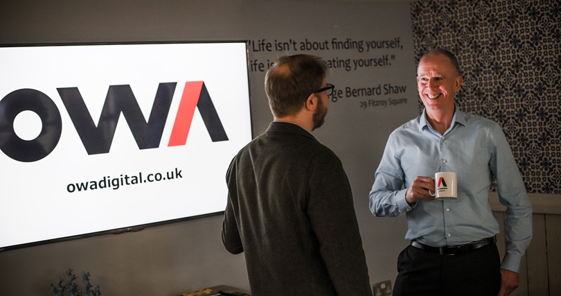 Two OWA team members in an office discussing legacy system support while drinking coffee together.