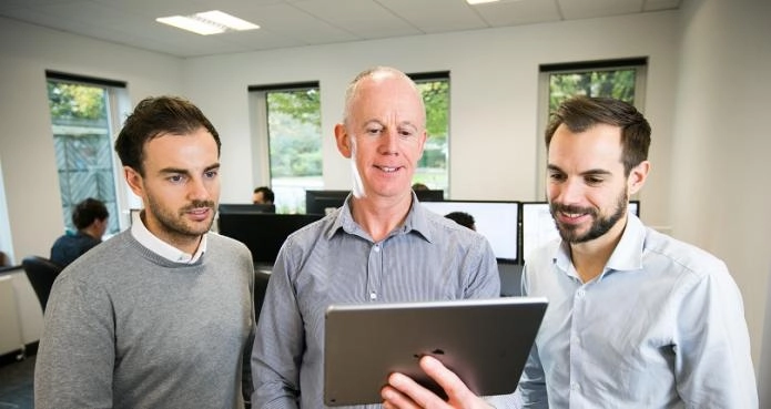 Three OWA team members in an office discussing Web Application Development while looking at a tablet together.