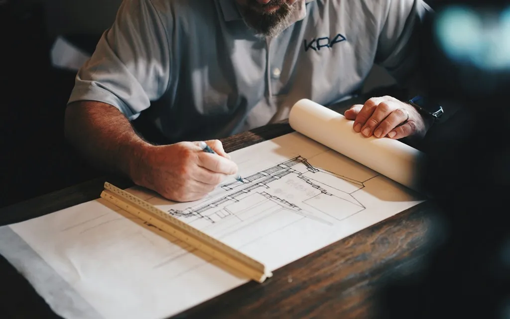 A man sits at a table, focused on working with a blueprint spread out in front of him.