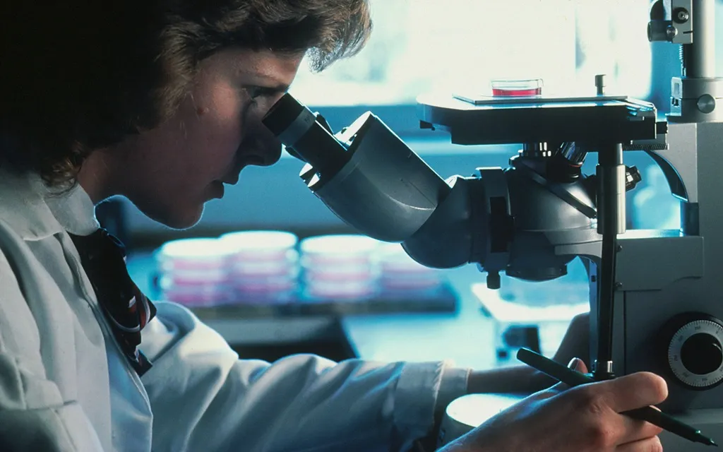 A woman in a lab coat examines a sample through a microscope in a laboratory setting.