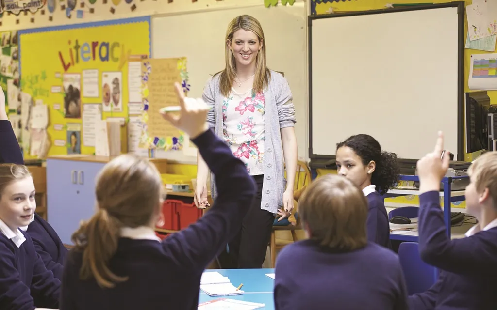 A teacher stands in a classroom as children eagerly raise their hands to answer questions.