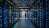A man stands in the center of a large server room filled with rows of servers and blinking lights.