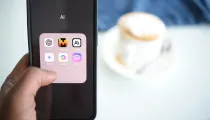 A hand holds a smartphone displaying a folder labeled "AI" with various app icons. A blurred cup of coffee and blue cloth are in the background on a table.